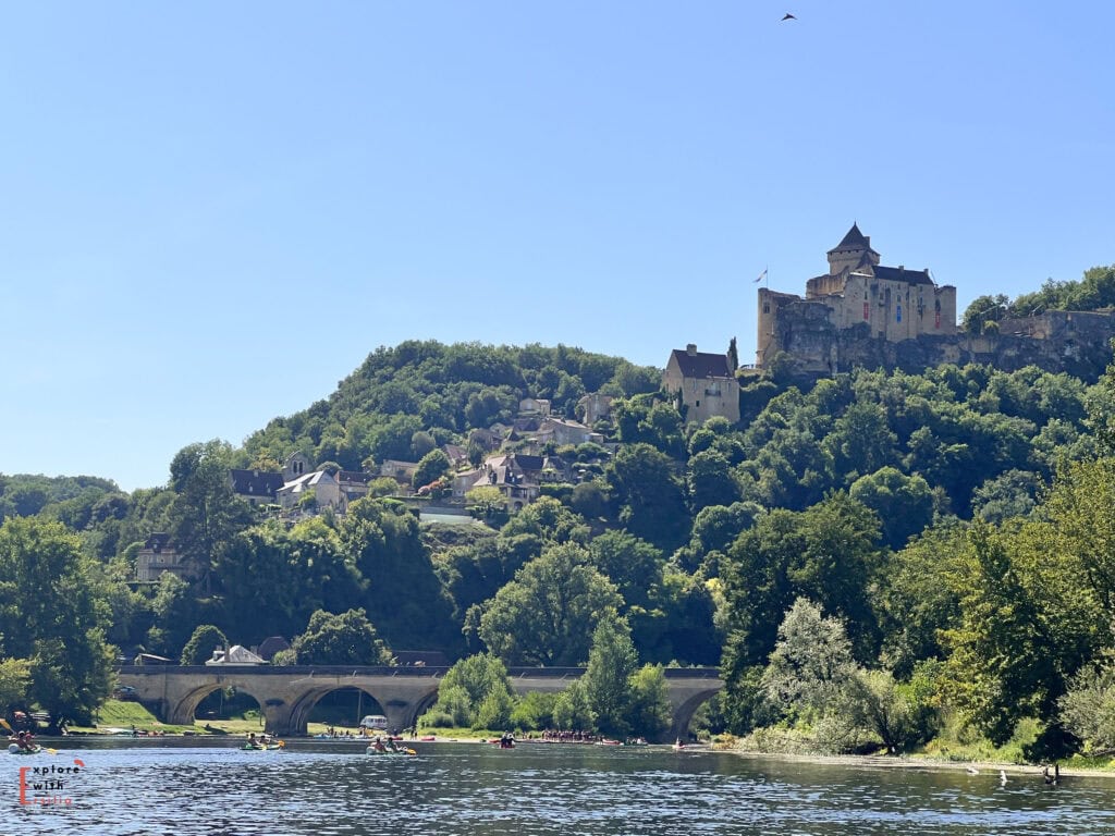 A view of Château de Castelnaud perched high on a hilltop above the Dordogne River on a bright summer day. Below, a stone bridge spans the river where numerous kayakers and canoeists gather near its arches. The medieval fortress stands prominently against a clear blue sky, with the village houses cascading down the forested hillside beneath it. The scene captures the popular river activities and historic architecture typical of the Dordogne Valley.