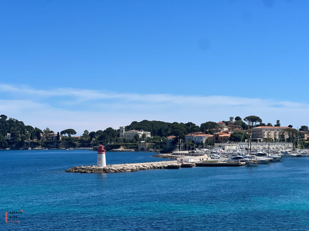 Wide coastal view in Saint Jean Cap Ferrat with clear turquoise water, rocky shoreline, and a sandy beach lined with people and boats under a bright blue sky.