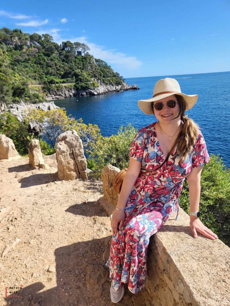 Woman in a floral dress and straw hat sitting on a stone wall in Saint Jean Cap Ferrat overlooking turquoise Mediterranean water and rugged coastal cliffs on a sunny day.