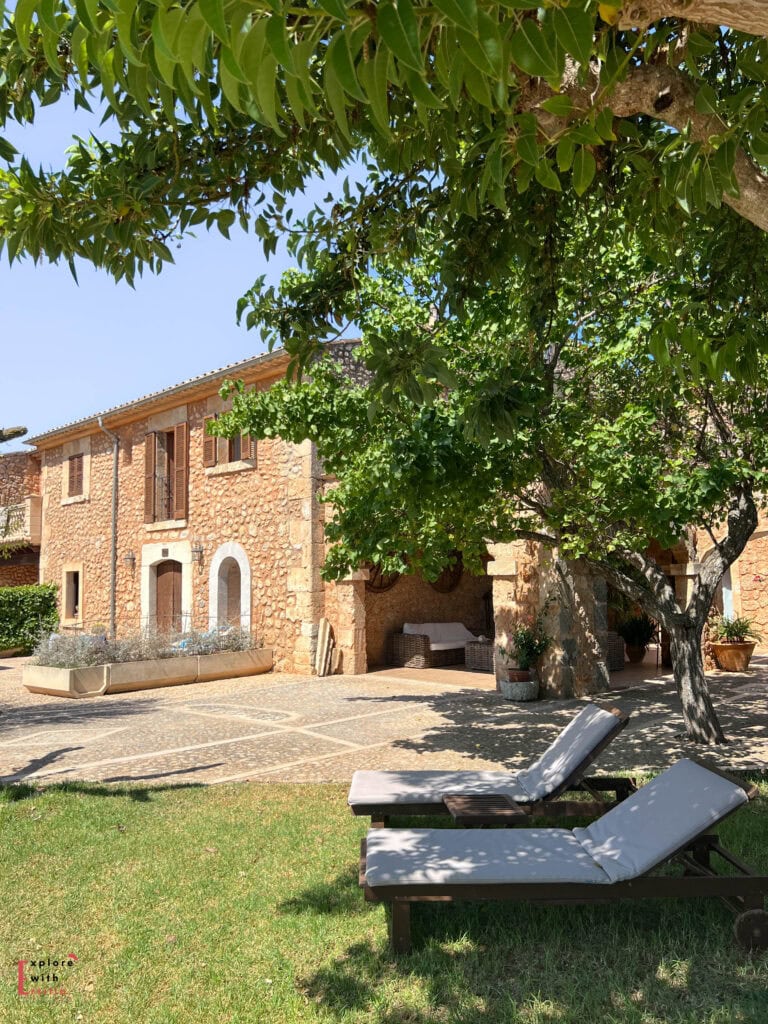 Traditional Mediterranean stone finca with light-colored loungers on a manicured lawn, shaded by lush green trees with an outdoor seating area and terrace under dappled sunlight, showcasing authentic Mallorcan architecture with wooden shutters and natural stone walls under blue sky.