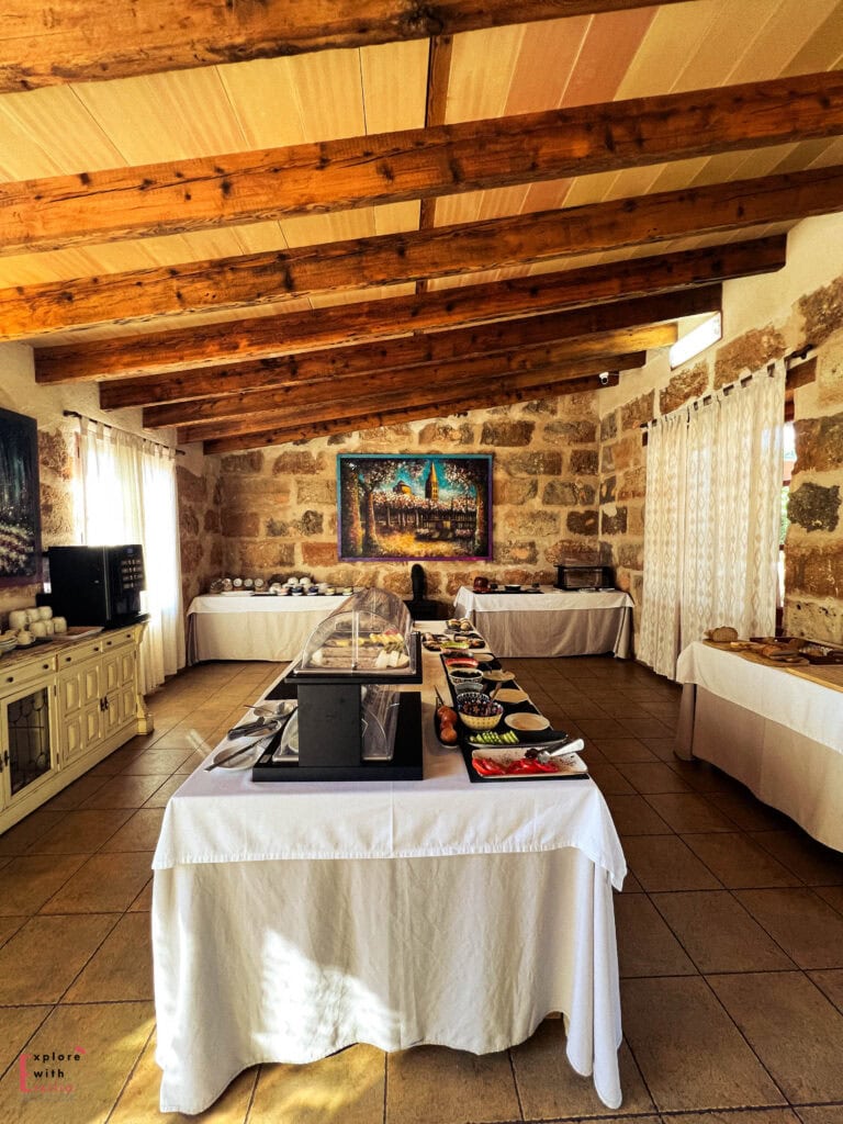 Rustic breakfast buffet room with exposed wooden beam ceiling, stone walls, and white-clothed tables arranged with various Mediterranean breakfast items, featuring a colorful painting on the wall and natural light filtering through linen curtains.
