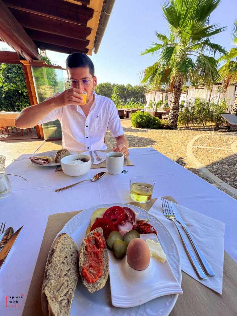 Person in white shirt and glasses enjoying an outdoor breakfast at a terrace table with traditional Mediterranean foods including bread with tomato spread, olives, eggs, and charcuterie. The background shows palm trees and a patio area with loungers on a stone-paved surface under clear blue sky.