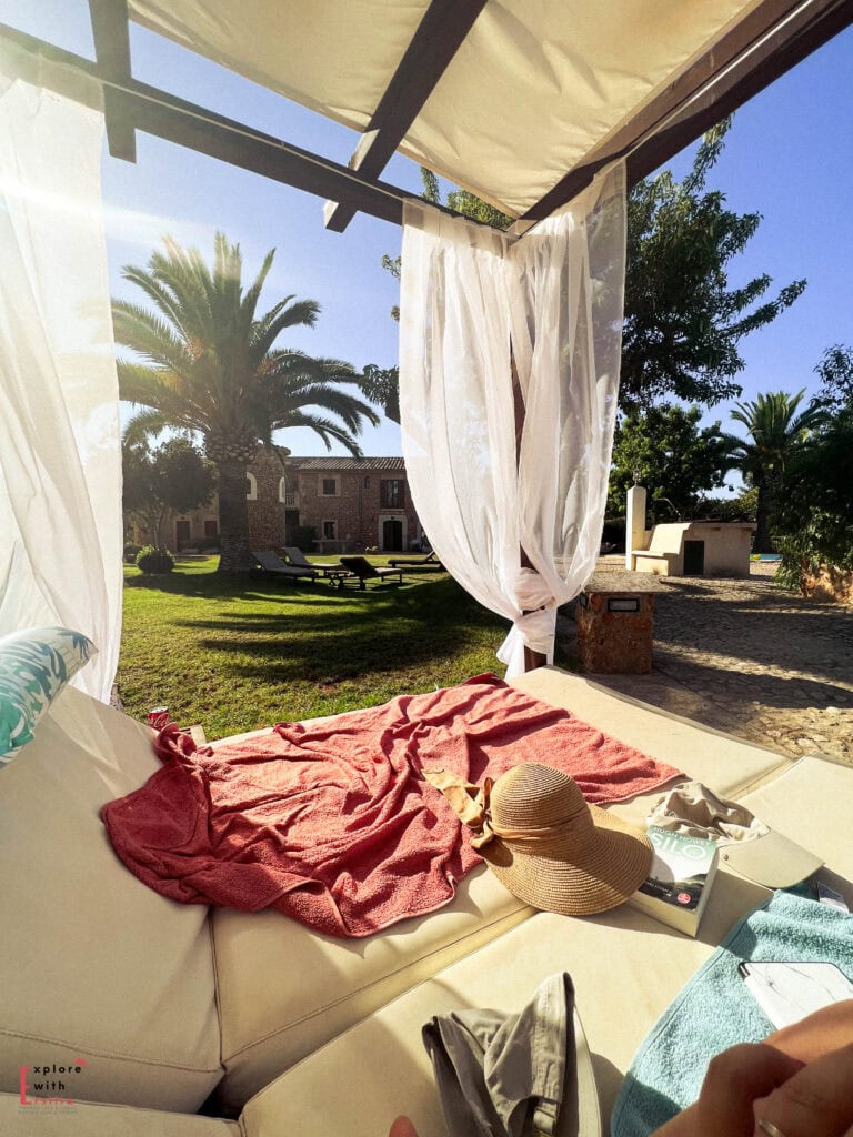 Luxurious canopy daybed with flowing white curtains, coral towel, and straw hat, overlooking a stunning Mallorcan estate with stone buildings, palm trees, and green lawns, creating a private oasis for relaxation under clear blue sky.