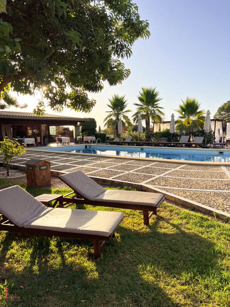 Luxury swimming pool area at sunset with turquoise water surrounded by stone patio, comfortable sun loungers with cushions on green lawn, palm trees, and an open dining pavilion with white-clothed tables in the background under clear blue skies.