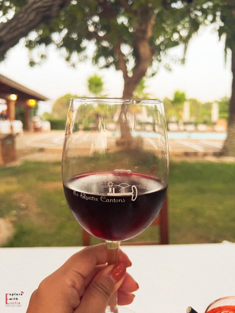 Hand holding a glass of red wine with "Es Quatre Cantons" engraved on it, in the foreground with a blurred swimming pool area, garden, and trees visible in the background during golden hour.