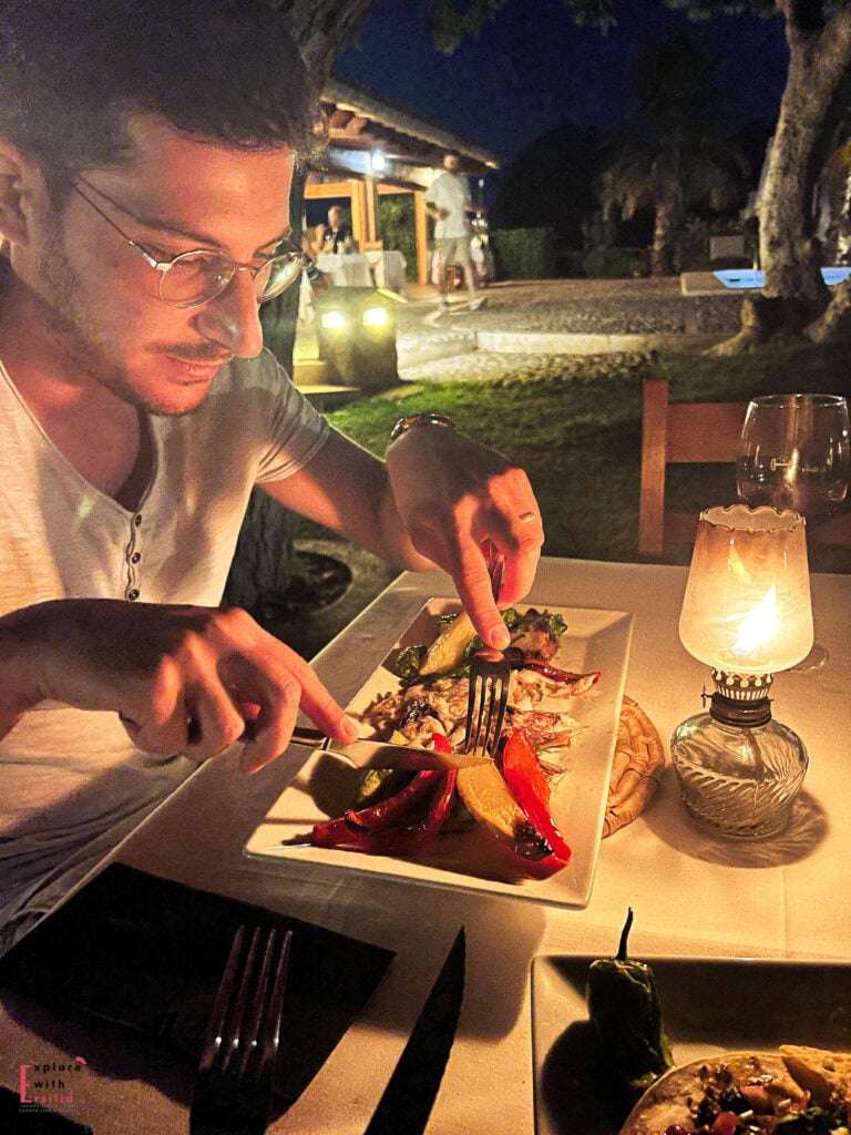 Person with glasses enjoying dinner by oil lamp light at an outdoor restaurant in the evening, using cutlery to savor a gourmet dish featuring roasted vegetables on a white square plate, with wine glasses visible on the table and soft lights illuminating the garden in the background.