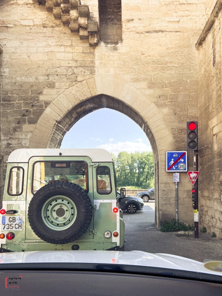 A vintage green Land Rover waits at a red light under a historic stone archway in Avignon, France. The medieval walls frame the narrow passage, blending centuries-old architecture with modern traffic signs.