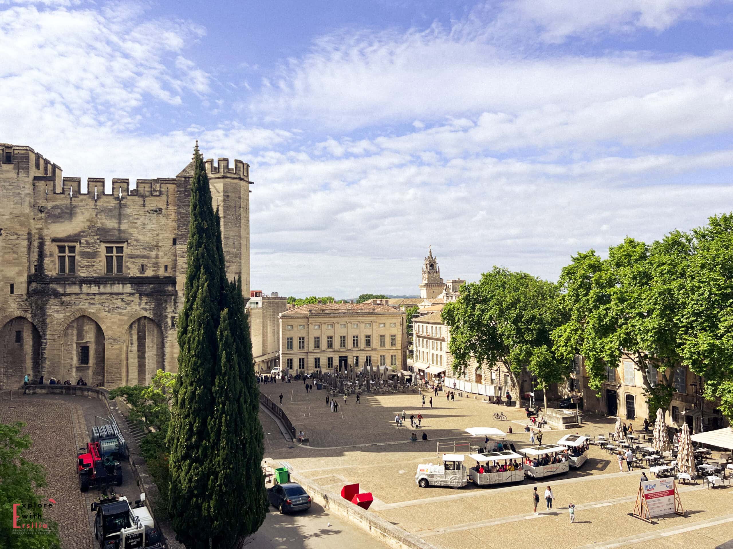 A wide view of Avignon’s historic square with the imposing stone façade of the Palais des Papes on the left and leafy trees lining café terraces on the right. Though busier and more urban, the mix of history and open space offers a cultural twist for honeymooners exploring Provence.