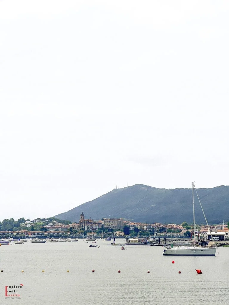 View across the Txingudi Bay from Hendaye, France, looking toward Hondarribia (Fuenterrabía) in Spain. The panorama shows sailboats moored in the calm harbor waters with the Spanish Basque town visible on the opposite shore. Traditional buildings with red-tiled roofs cluster around a church with a prominent bell tower, all nestled at the foot of Mount Jaizkibel. This scene captures the transboundary nature of the Basque Country where France meets Spain across the bay.