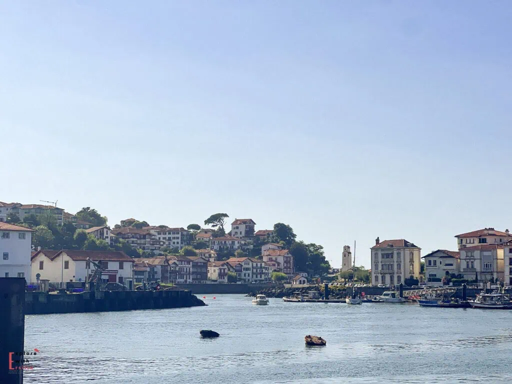 Panoramic harbor view of Saint-Jean-de-Luz, a picturesque fishing port and resort town in the French Basque Country. The image shows the town's characteristic white buildings with red roofs cascading down the hillside around the protected bay. Small fishing boats and pleasure craft are moored in the calm harbor waters, with the historic town center visible on the right featuring traditional Basque architecture, and a church tower (likely the Church of Saint John Baptist) rising among the buildings under a clear blue sky.