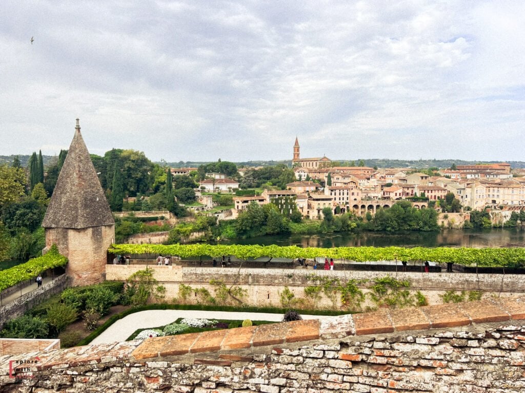 Panoramic view of Albi in summer, with historic brick buildings, arched riverside houses, and a church tower rising above the Tarn River. A shaded riverside walkway and lush greenery highlight why Albi is an appealing summer destination in southern France, blending history with relaxed outdoor scenery.