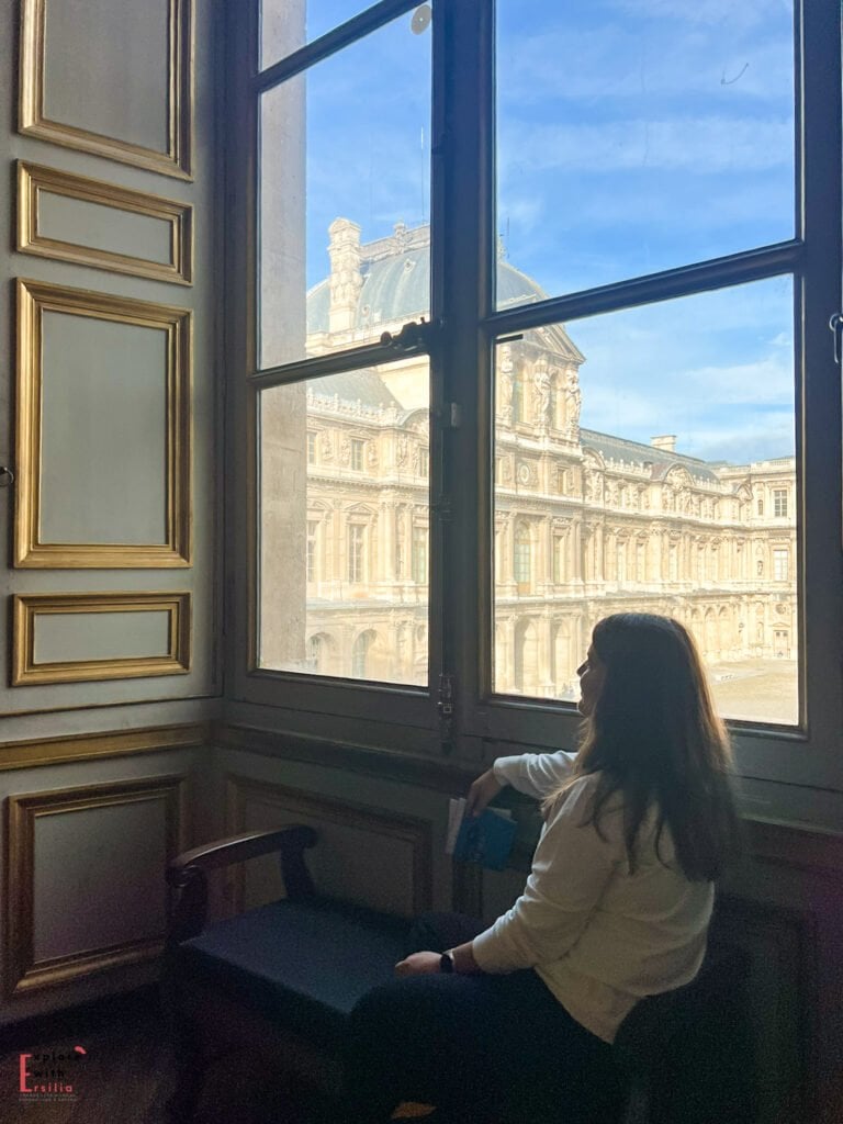 A woman sitting by a grand window inside the Louvre, gazing out at the ornate palace architecture bathed in natural light, with gilded wall details framing the quiet moment.