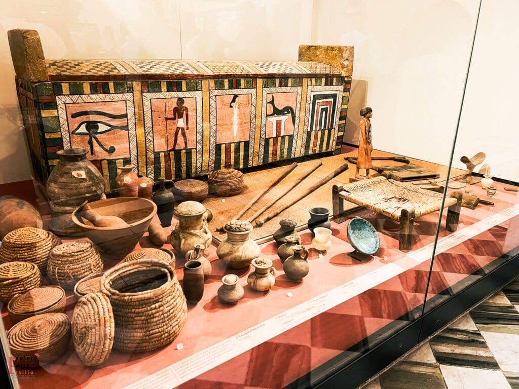 A painted wooden Egyptian box decorated with hieroglyphs and symbols, surrounded by pottery, tools, and miniature figures from daily life in ancient Egypt.