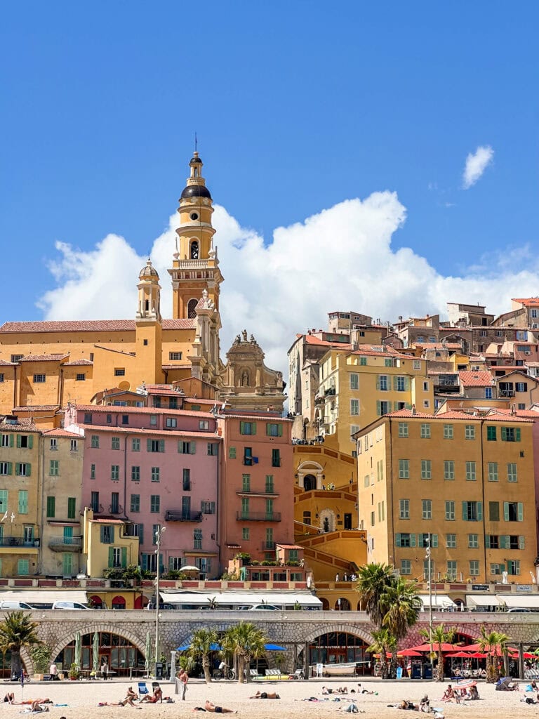 Vibrant hillside view of Menton, France, showcasing rows of pastel-colored buildings stacked along narrow streets with the striking bell tower of the Basilica of Saint-Michel-Archange rising above. The scene is bathed in sunlight under a bright blue sky with fluffy clouds.