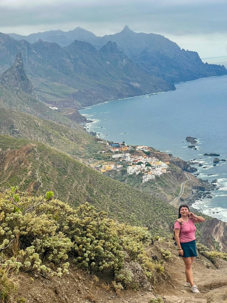 A woman stands on a lush mountain trail overlooking the dramatic northern coastline of Tenerife, with steep cliffs, a winding road, and the village near Playa de Benijo visible far below along the rocky shore.