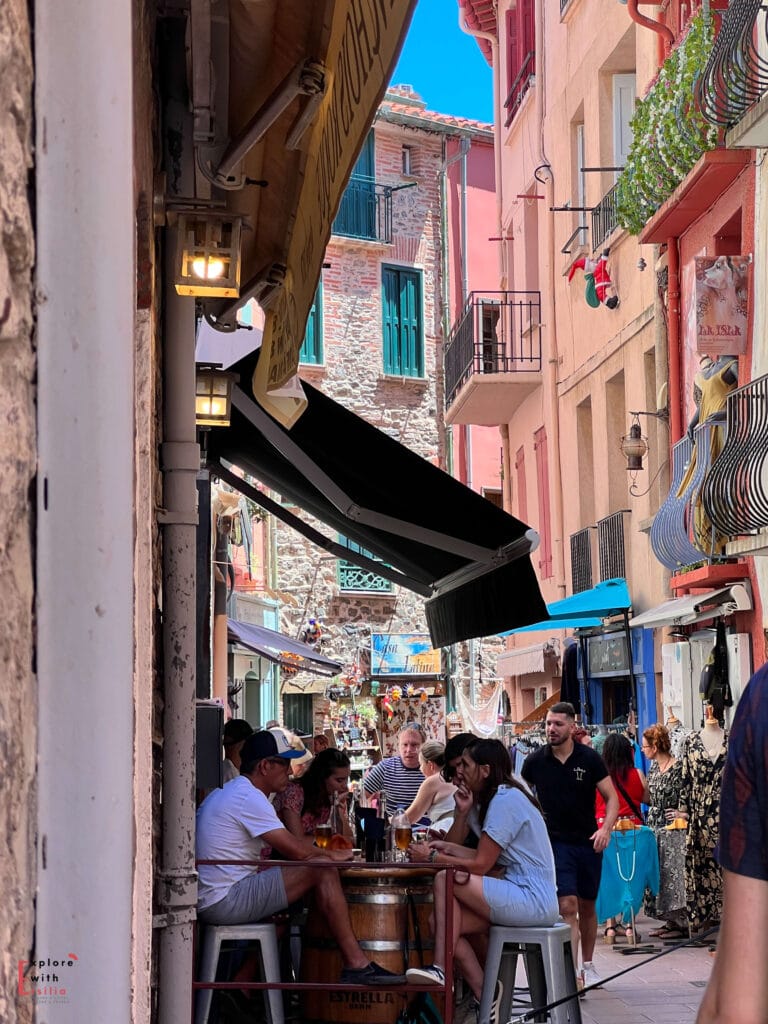 Bustling narrow street in Collioure with people dining at small café tables beneath awnings, surrounded by pastel buildings and balconies. The lively alley captures everyday life in this coastal town and beach town, showing the charm that makes Collioure a hidden gem town in France.