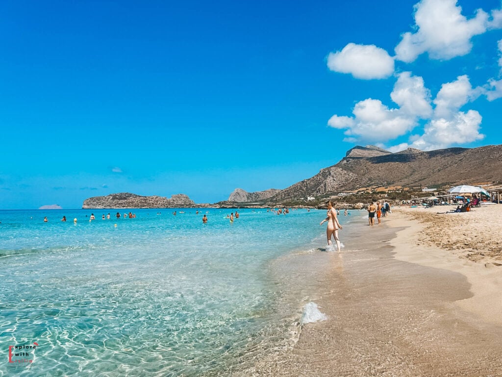 Wide stretch of Falasarna Beach in Crete with golden sand meeting crystal clear turquoise water. Swimmers dot the calm sea while rugged mountains rise in the background under a bright blue sky, capturing the wild beauty and open space that make this beach one of Crete’s most famous coastal escapes.