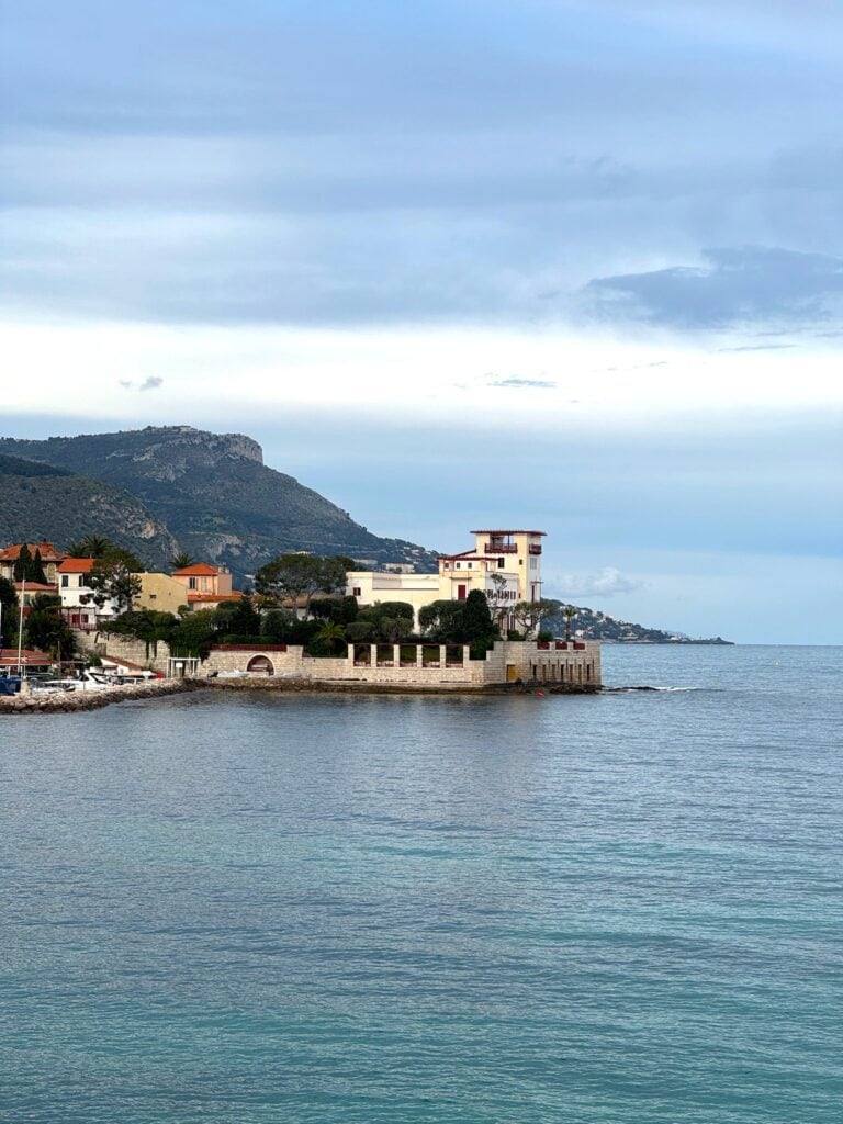 Villa Kérylos in Saint Jean Cap Ferrat overlooking calm Mediterranean water, with a pale waterfront mansion, stone terrace, and mountains in the background under a