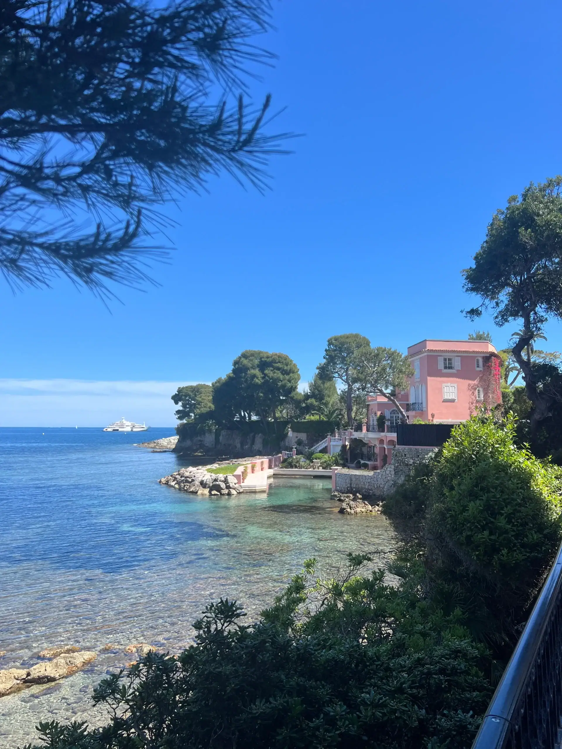 Coastal view in Saint Jean Cap Ferrat with a pink seaside villa, clear turquoise water, rocky shoreline, and a yacht in the distance under a bright blue sky.