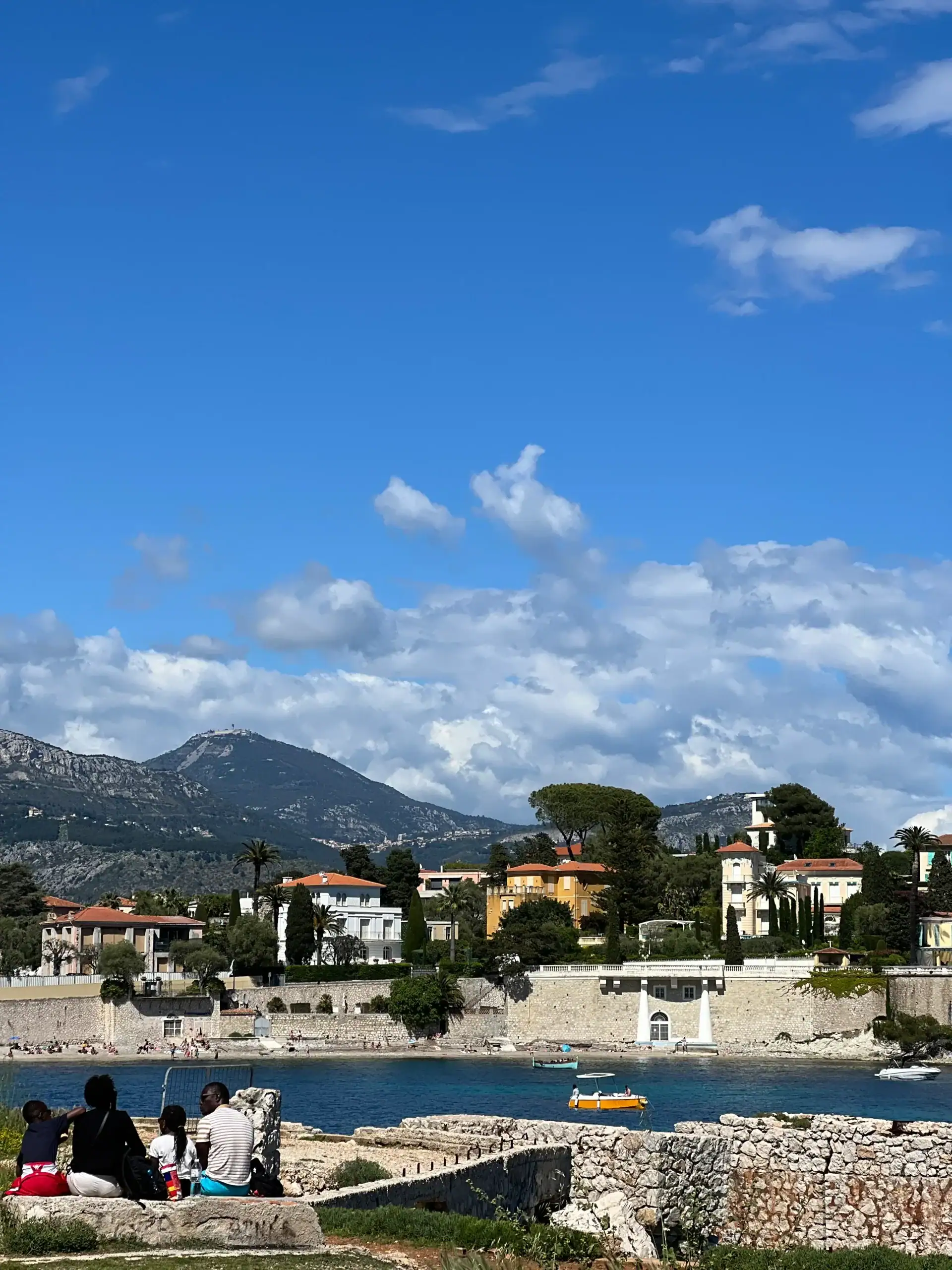 View in Saint Jean Cap Ferrat with people sitting by the water, colorful houses along the shoreline, and mountains rising in the background under a partly cloudy sky.