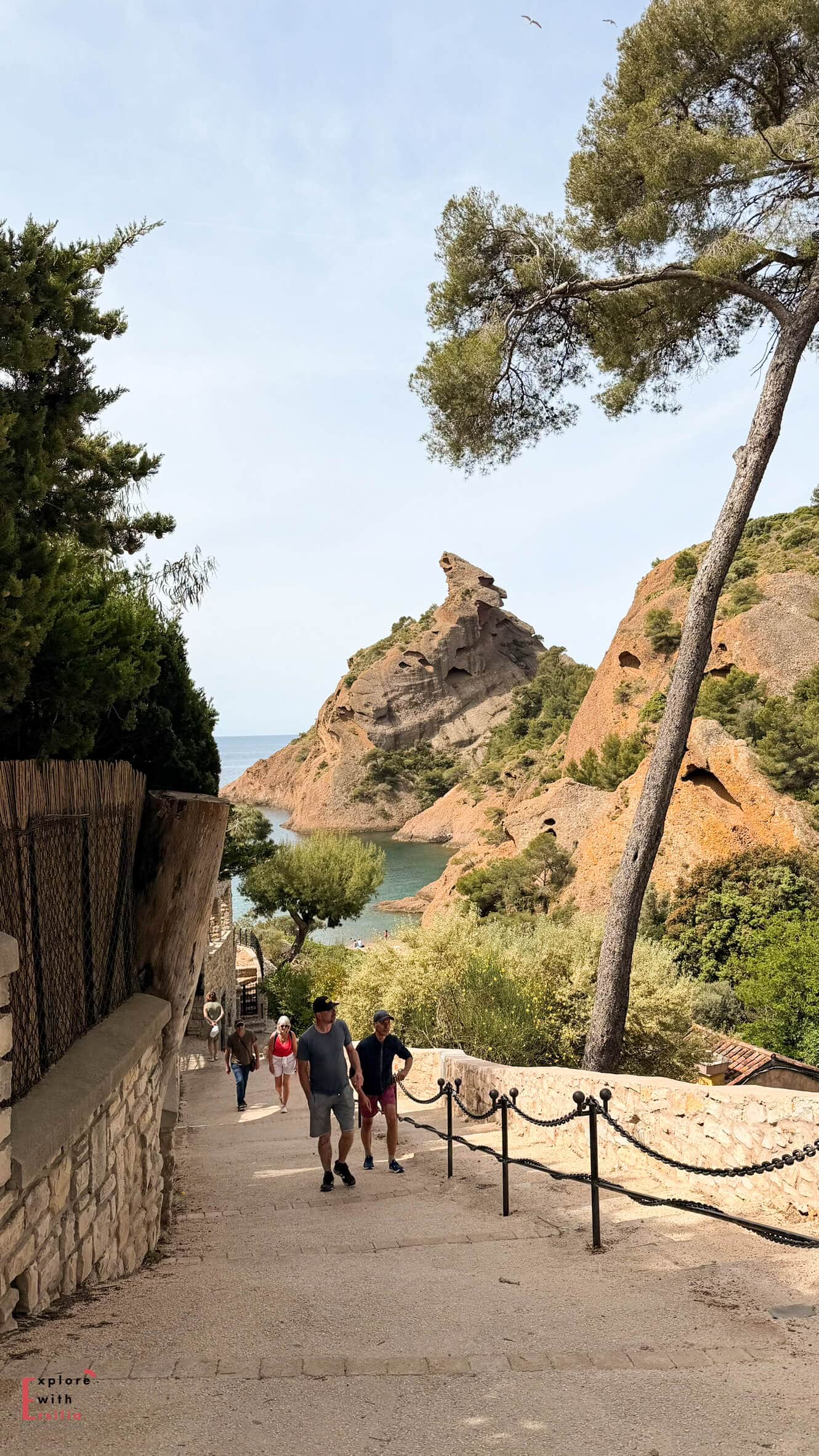 Visitors walking down a sloping path toward Calanque Figuerolles with orange rock formations and Mediterranean views ahead.