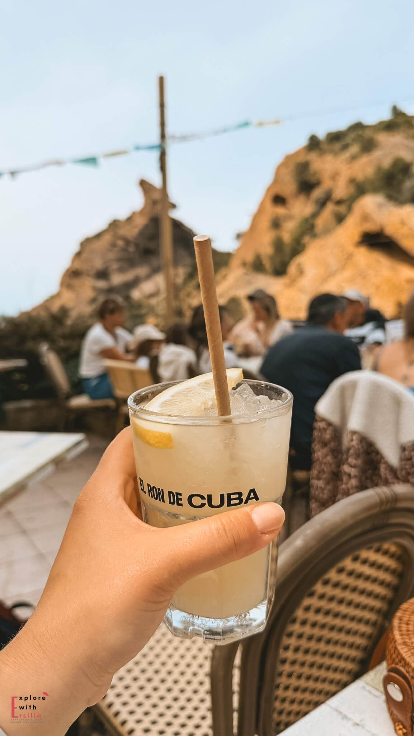 Hand holding a cold drink on an outdoor terrace overlooking the cliffs and coastline at Calanque Figuerolles.