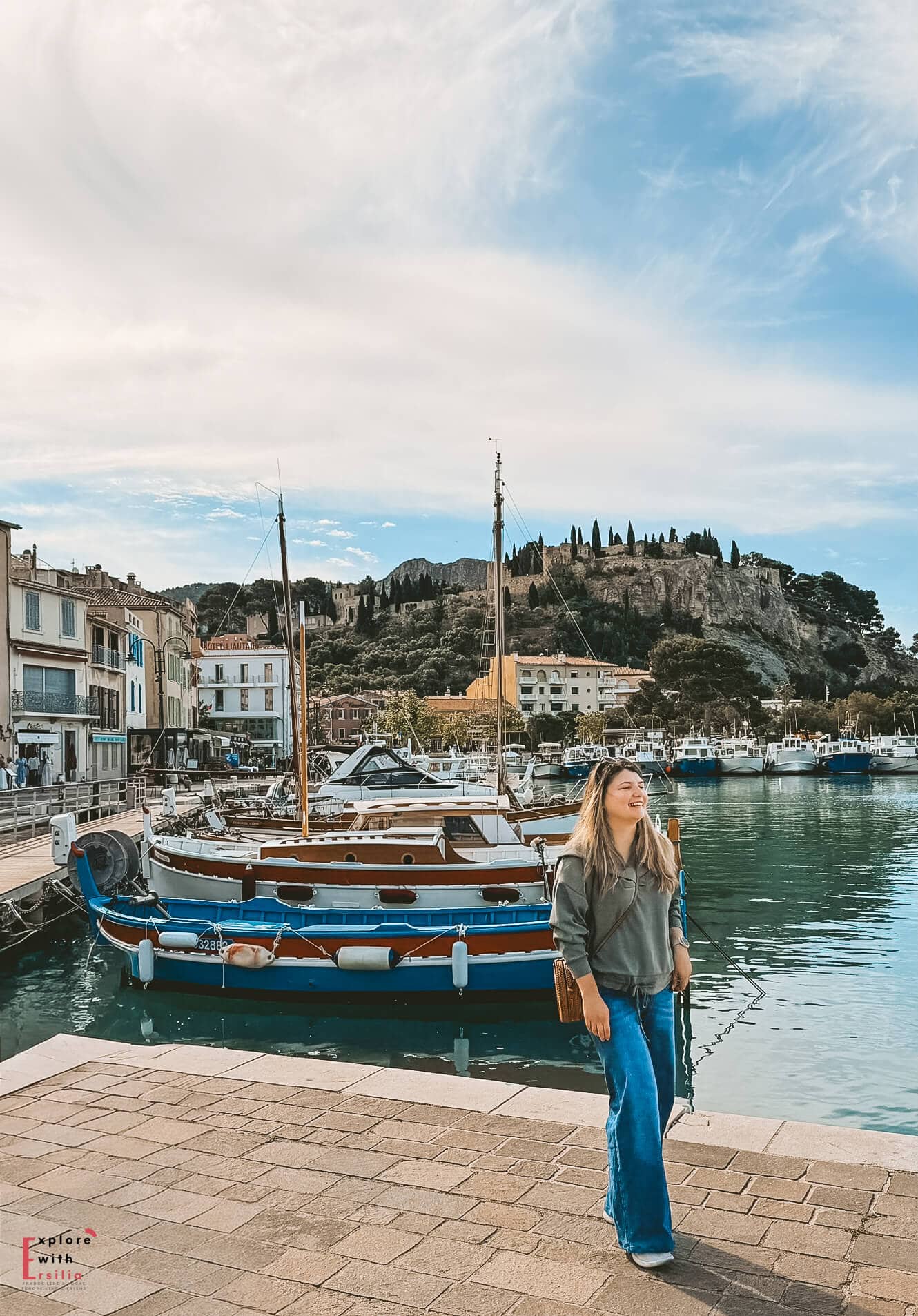 Woman (me) standing beside traditional boats on the Cassis waterfront with calm harbor water and hillside views behind her.