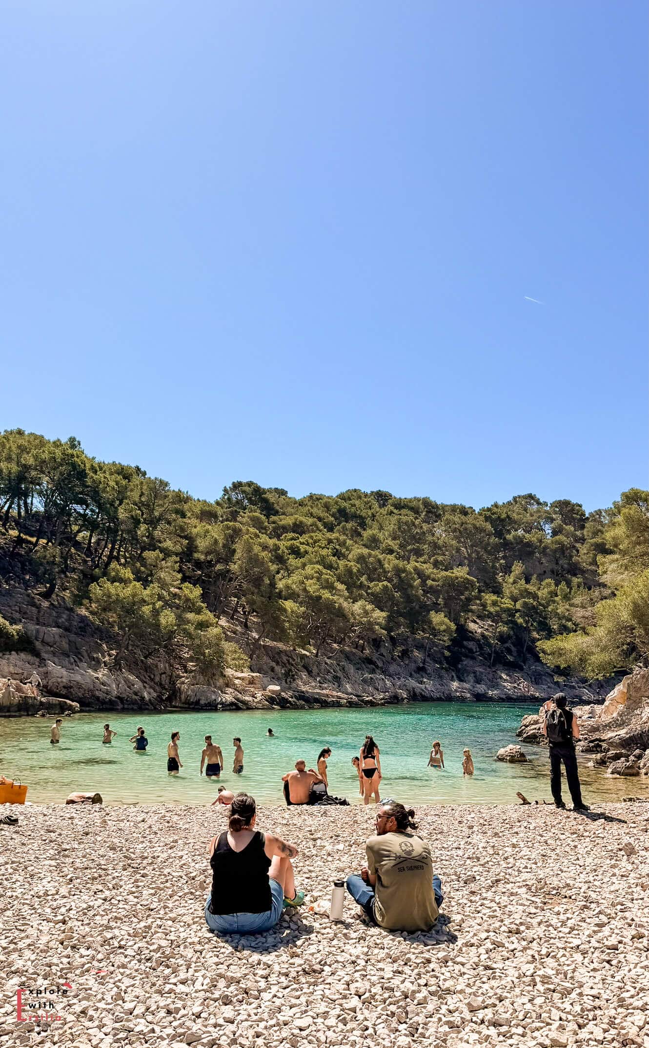 Pebble beach at Port Miou with visitors relaxing beside clear turquoise water surrounded by pine-covered hills.
