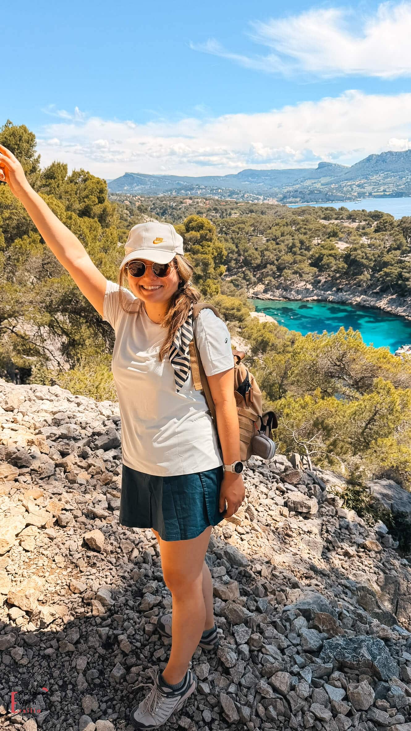 Woman (that's me!) standing on a rocky overlook above the Calanques with turquoise coves, pine-covered hills, and the Mediterranean Sea behind her.