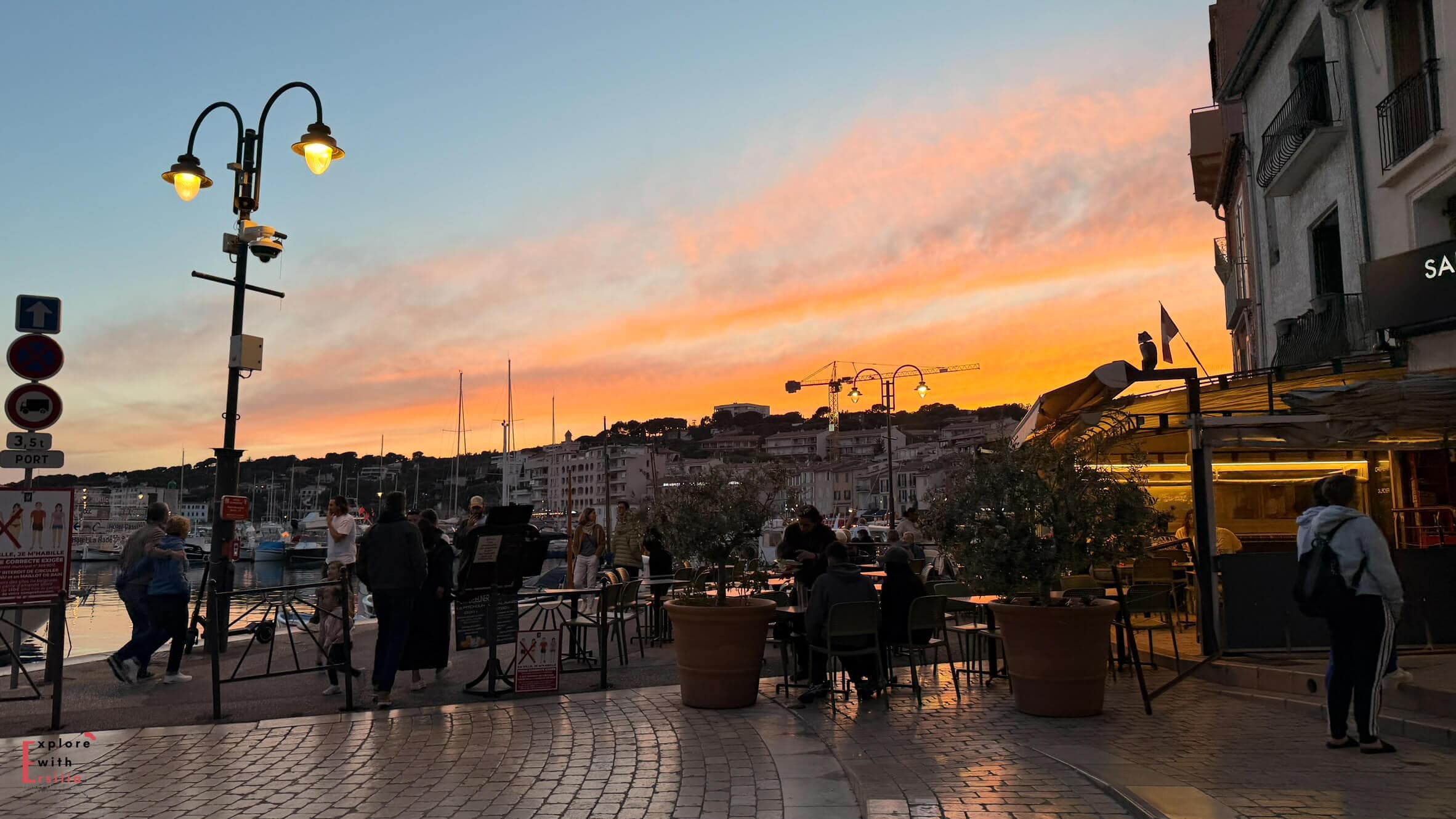 Sunset over Cassis harbor with people gathering along the waterfront and warm evening light reflecting across the sky.
