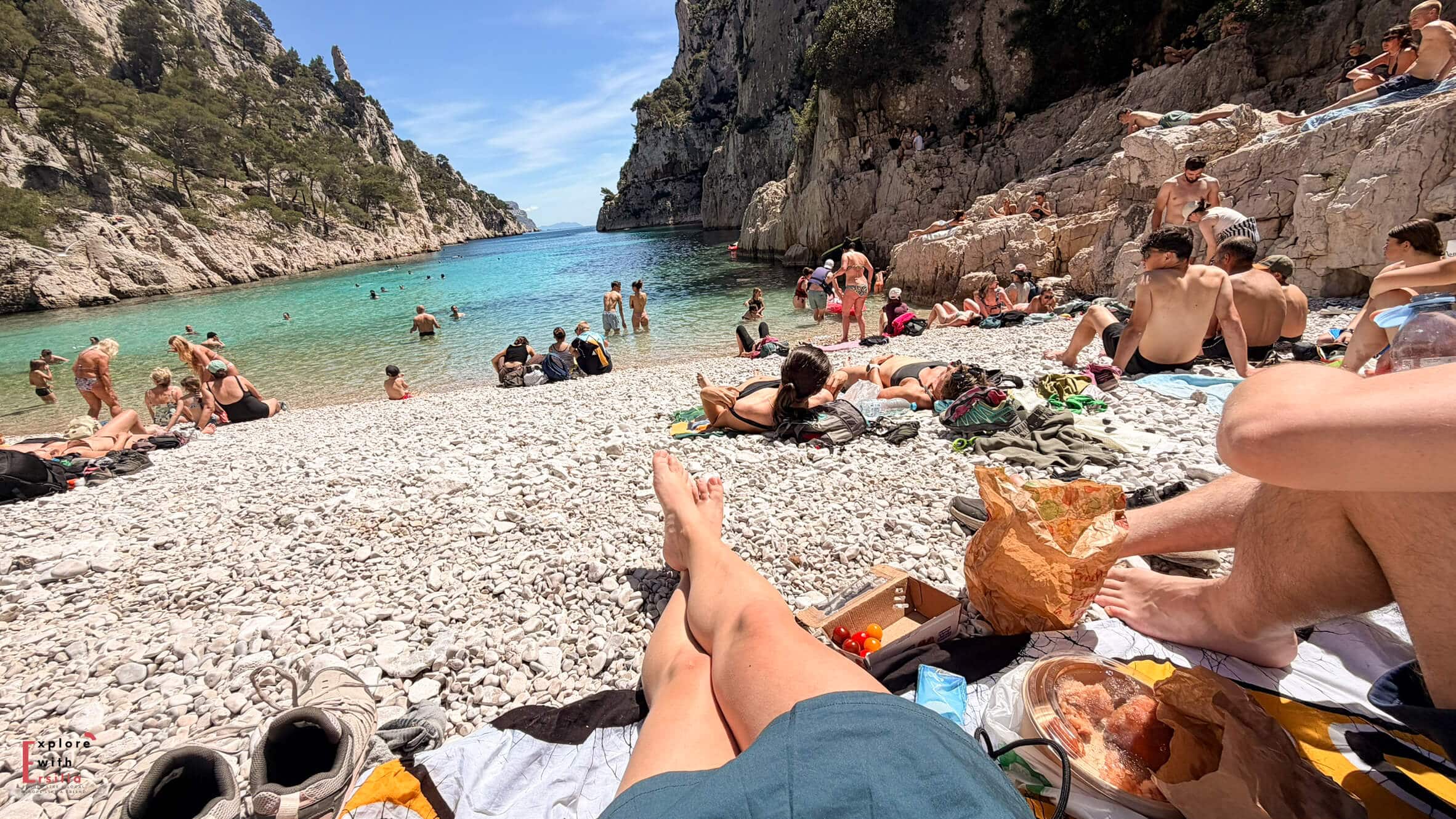 two persons having a picnic on the peblle beach with many other people around, showing it is crowded