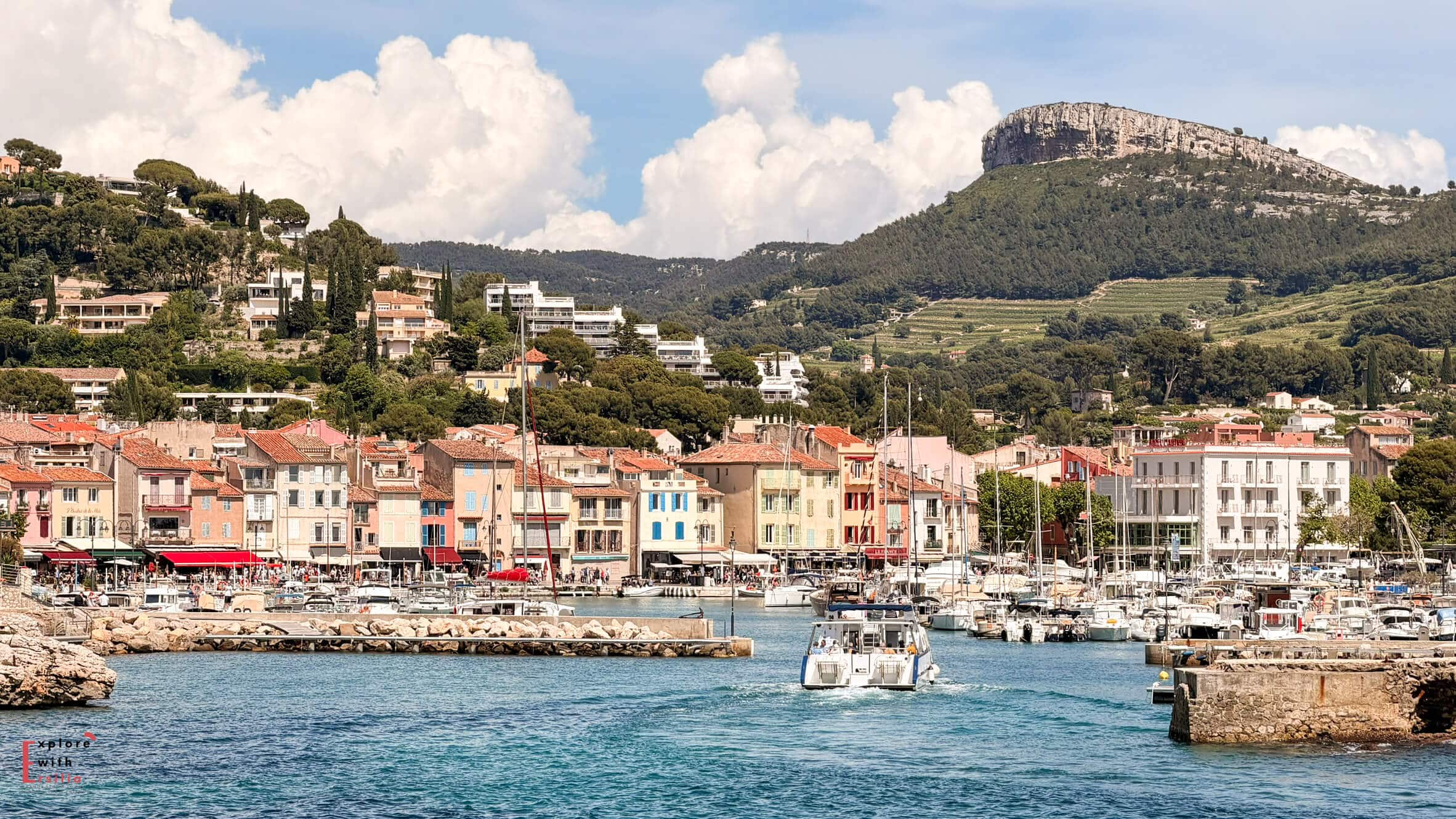 View across Cassis harbor with fishing boats, waterfront buildings, and green hills rising behind the town.