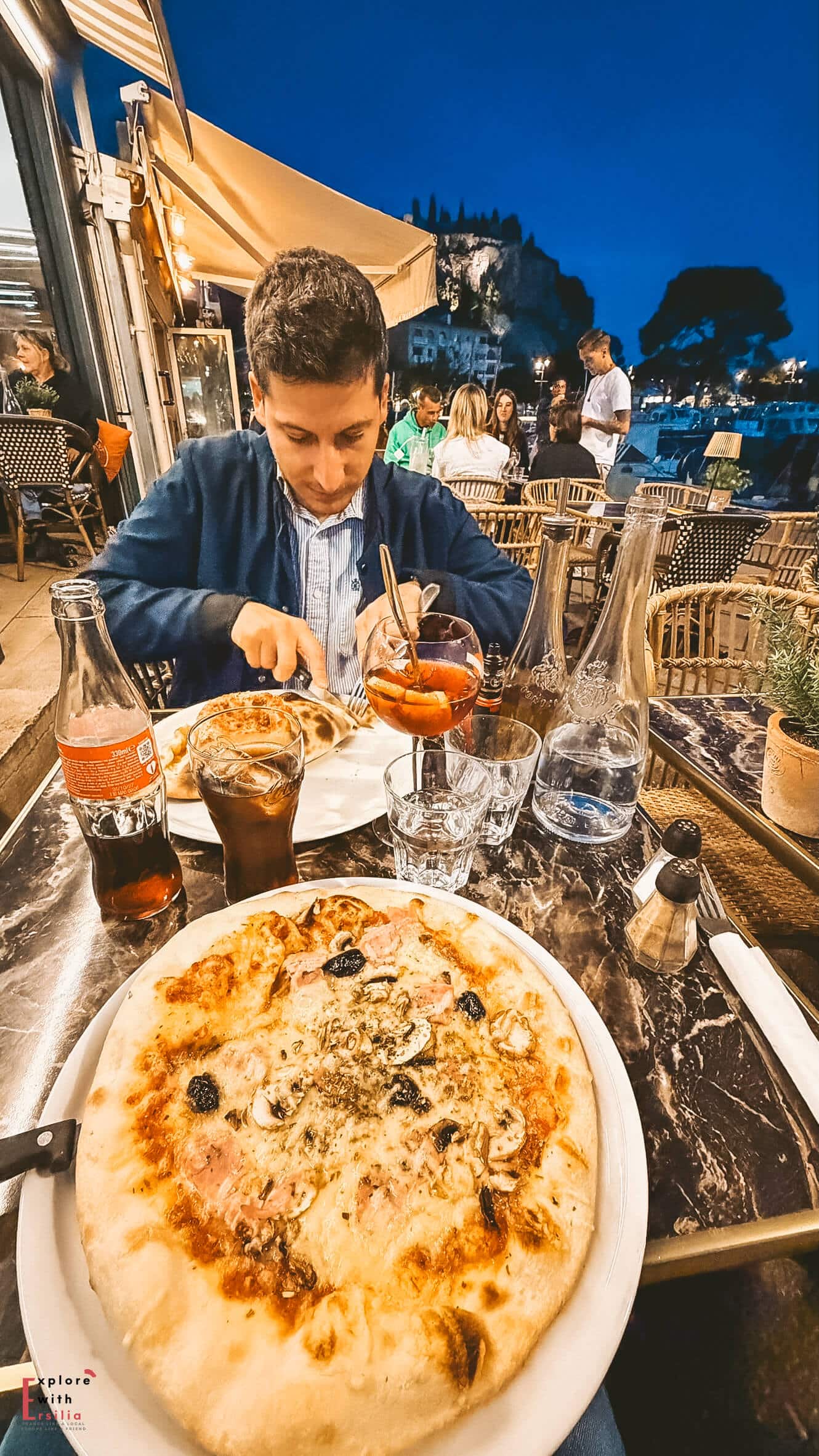 Outdoor dining terrace at Le 8 et Demi overlooking the lively streets of Cassis at sunset. Wood-fired pizza served on an outdoor restaurant table with drinks and evening dining atmosphere in Cassis.