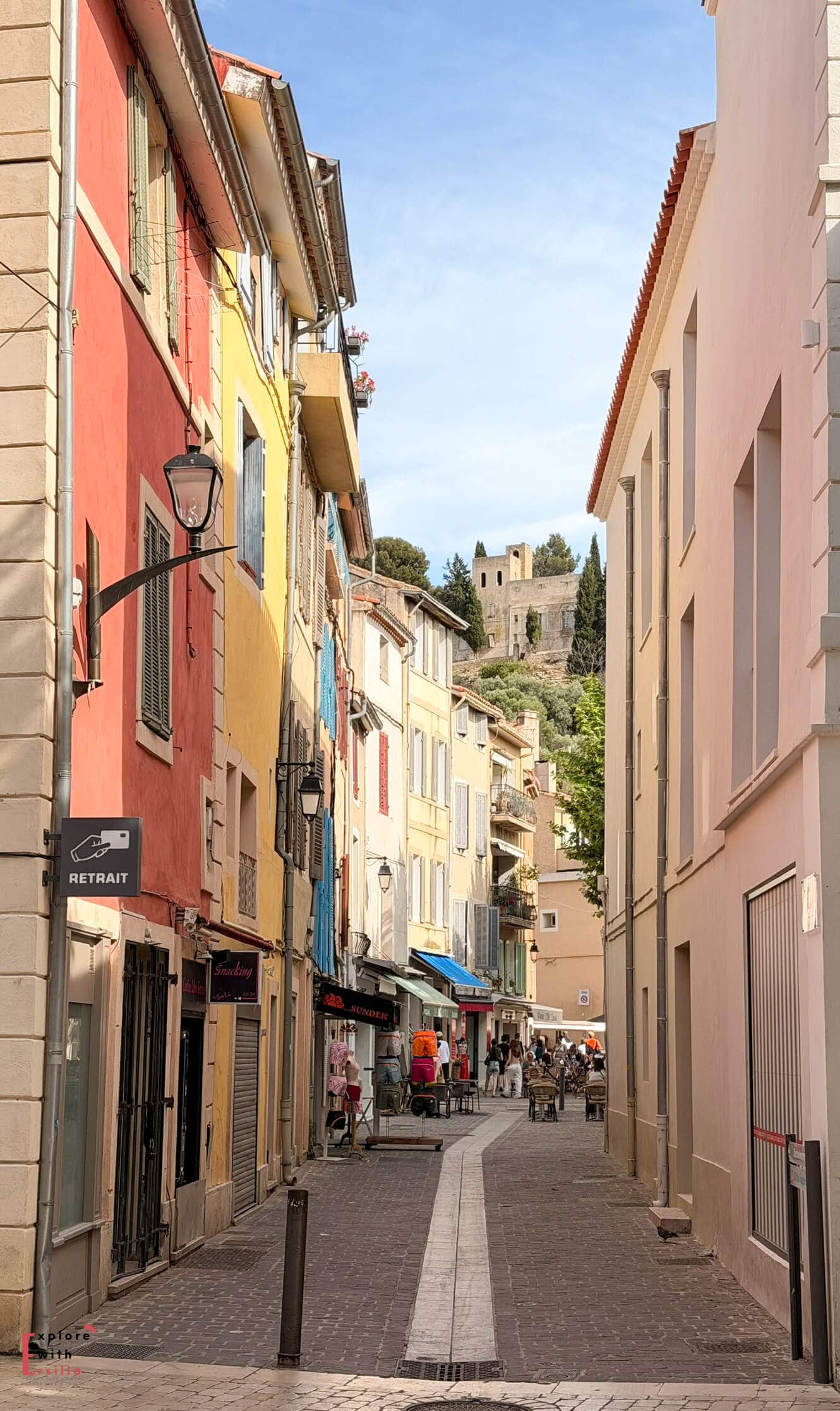 Narrow street in Cassis lined with pastel-colored buildings leading toward a hilltop view at the end of the lane.