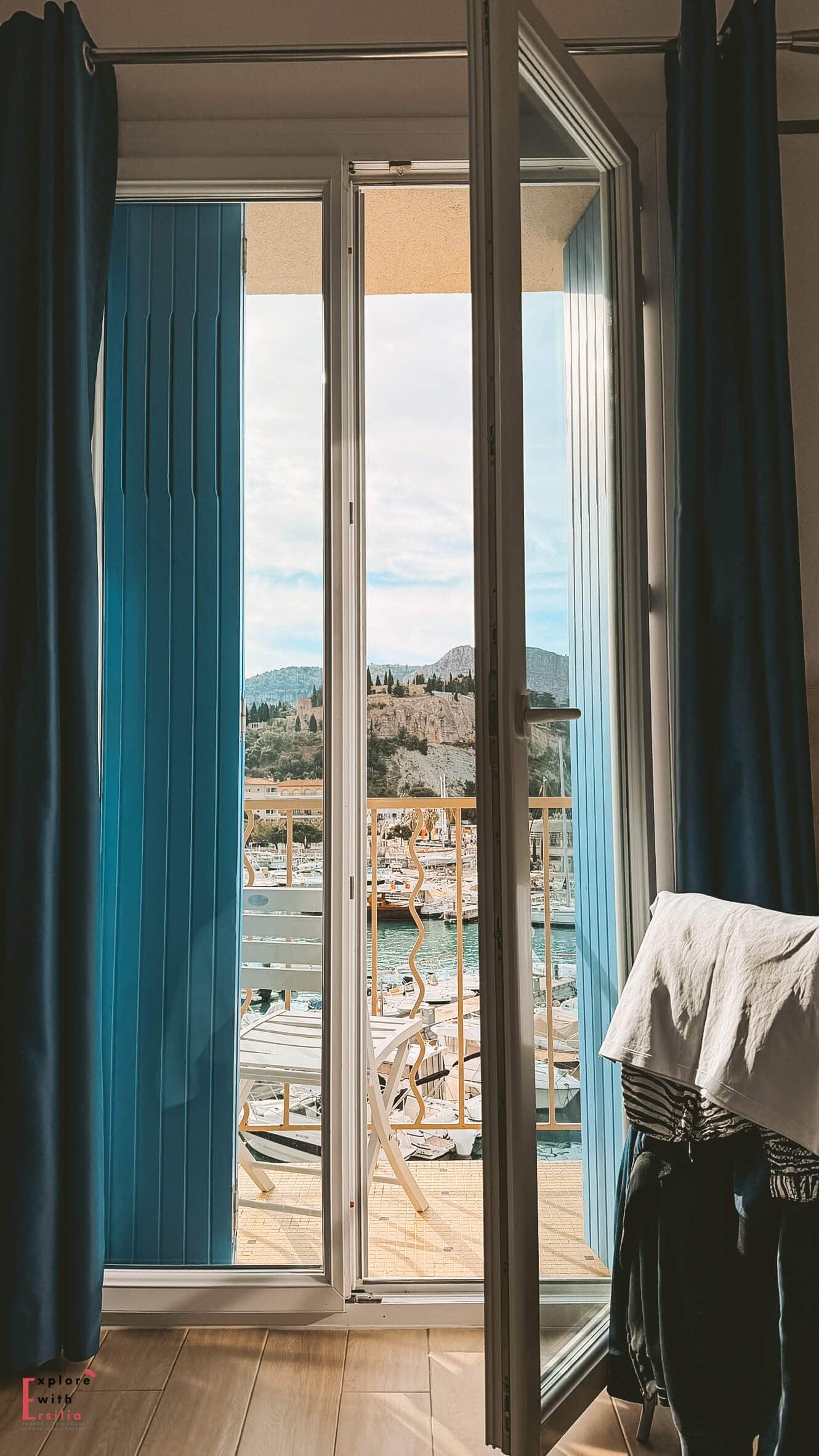 View from a hotel room window looking out over the Cassis harbor and waterfront through open glass doors framed by blue curtains.