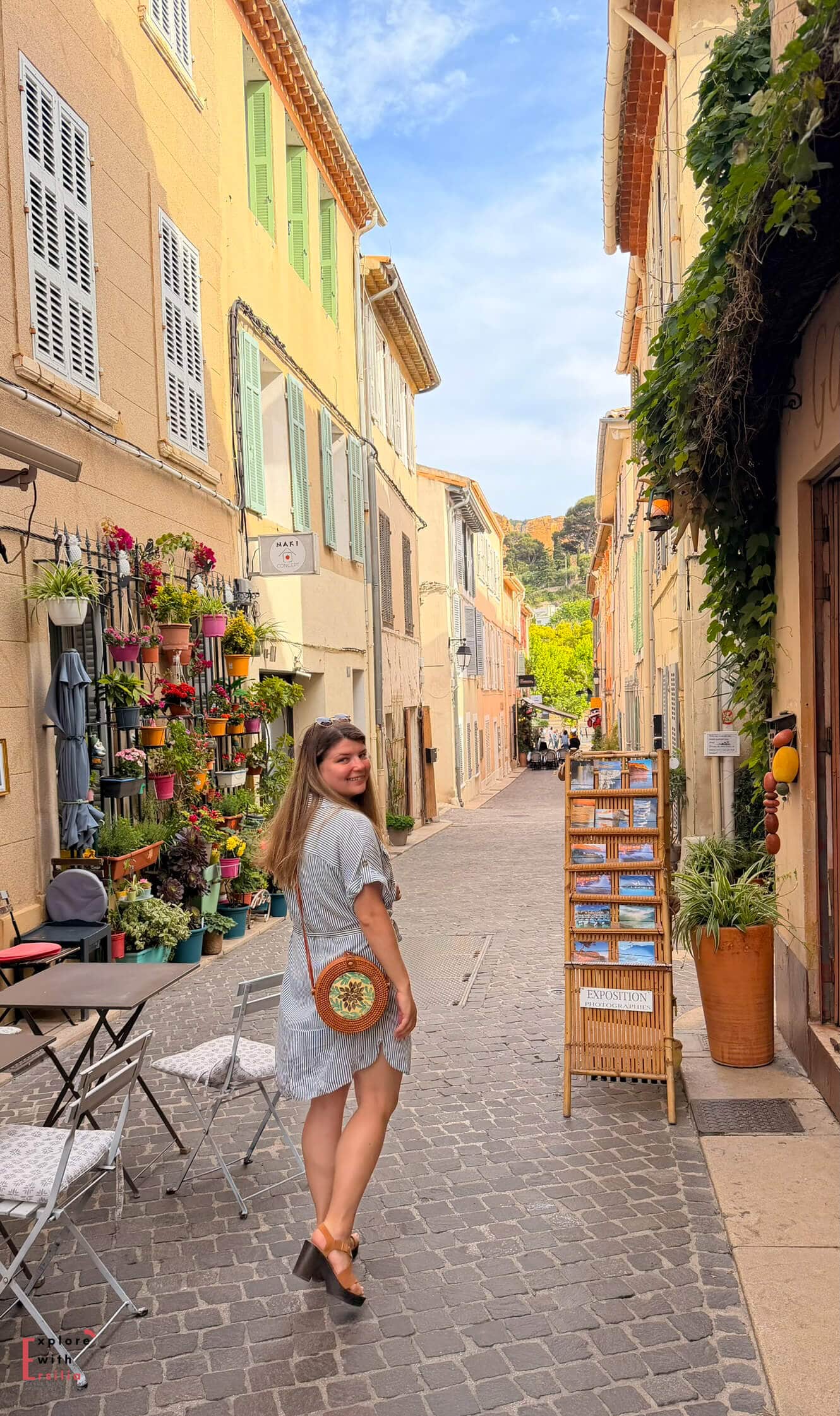 Woman walking through a colorful street in Cassis past cafés, flower displays, and pastel village buildings.