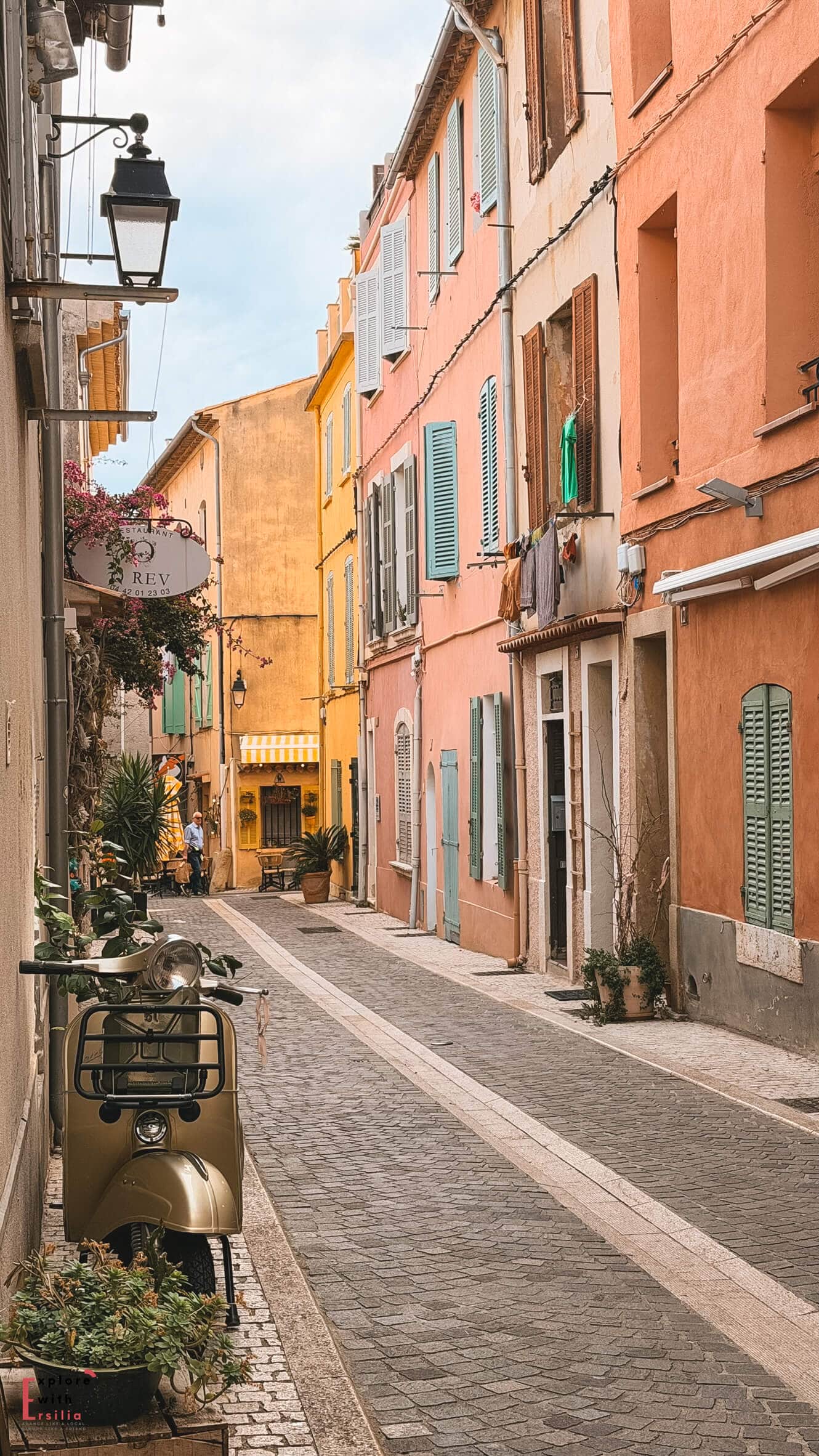 Quiet cobblestone street in Cassis bordered by peach and pink façades with shuttered windows and outdoor seating.