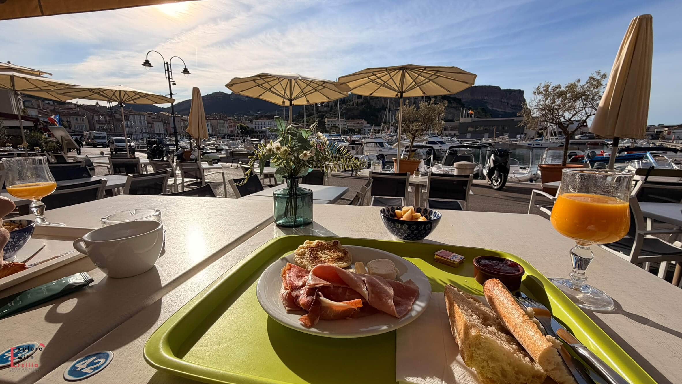 Breakfast terrace overlooking Cassis harbor with boats, waterfront buildings, and limestone cliffs illuminated by morning light.