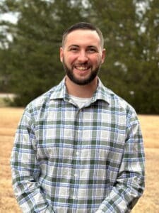 Pastor Brandon Deas standing in front of a row of pine trees.