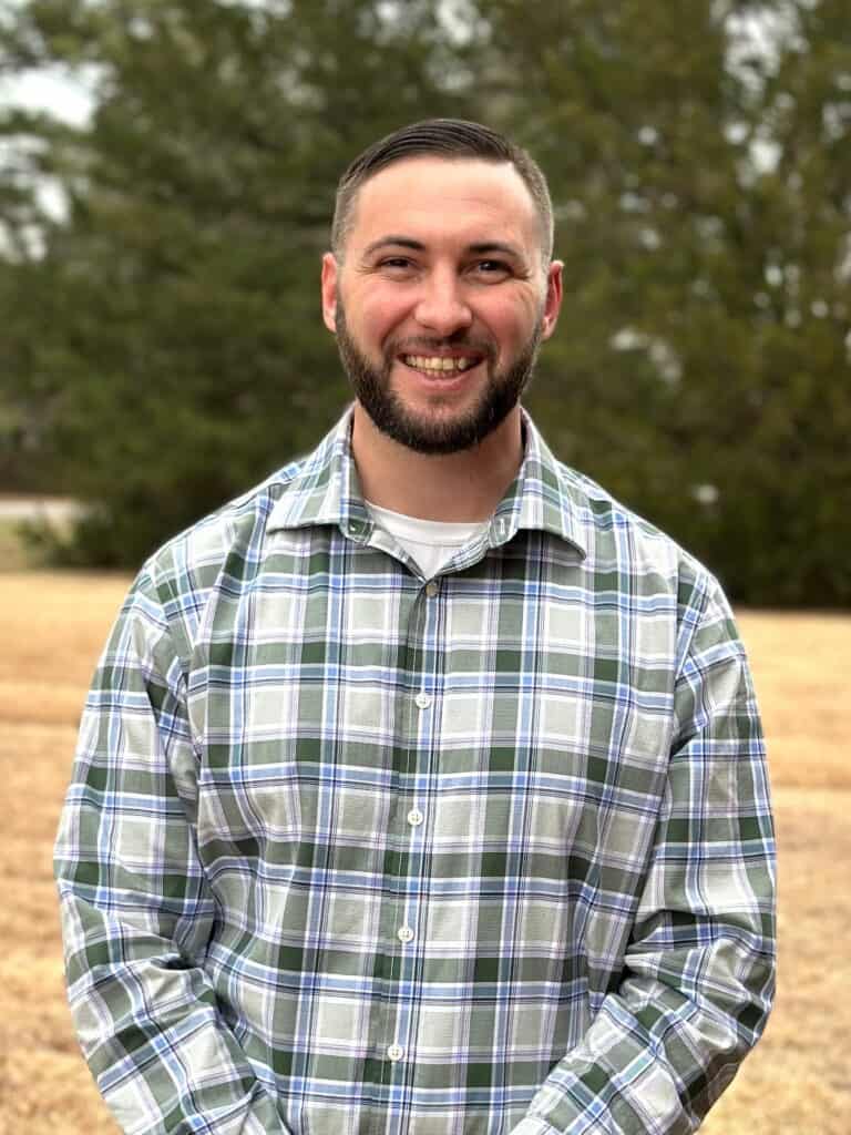 Pastor Brandon Deas standing in front of a row of pine trees.