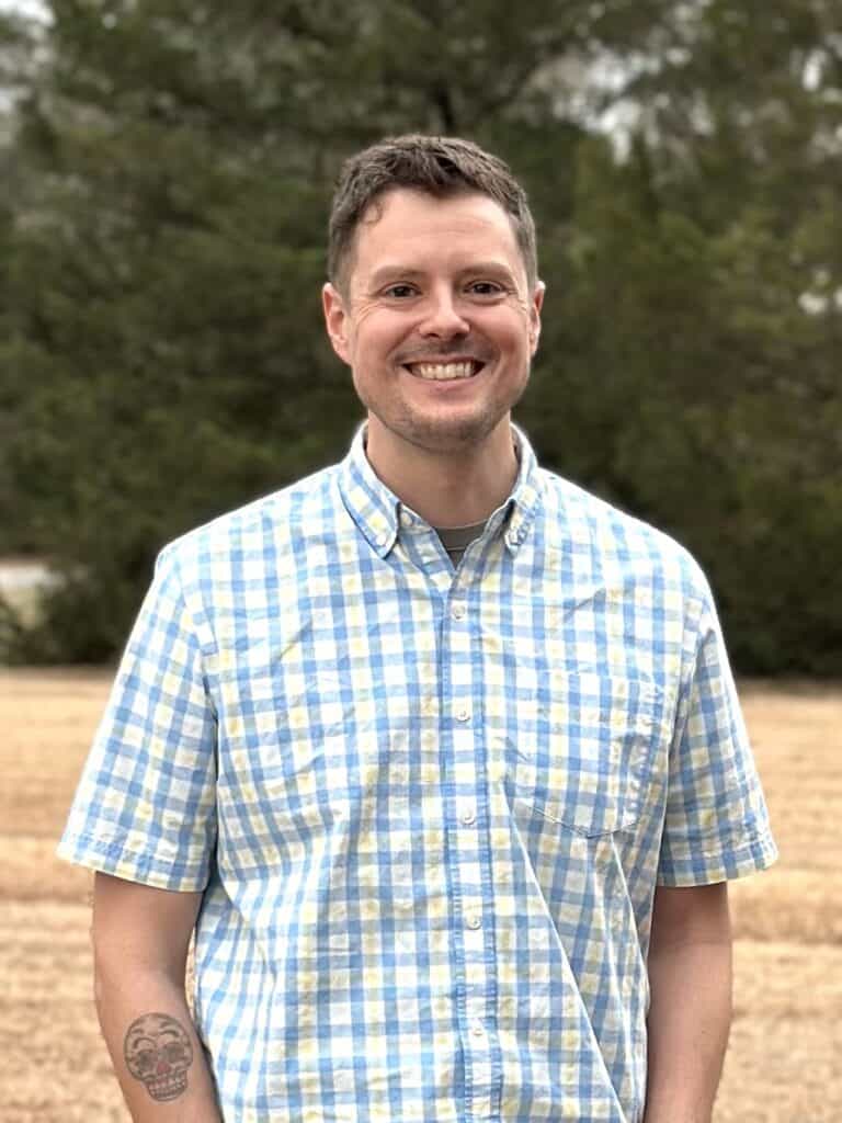 Headshot of Casey Robinson. He is standing in front of a row of pines.