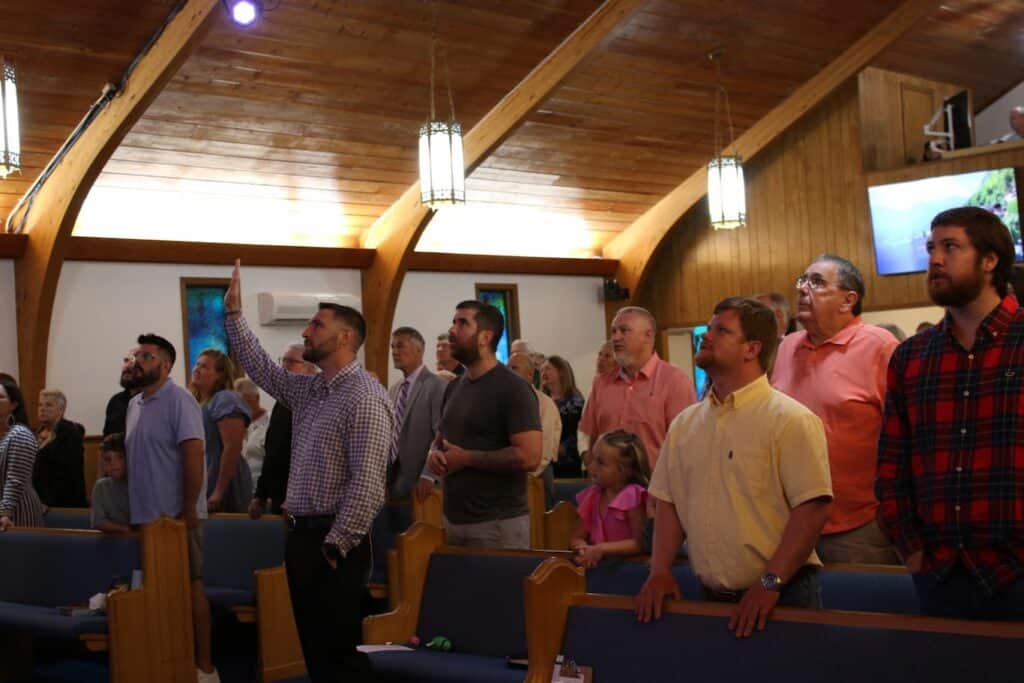 Inside of the Sanctuary. People are standing and singing. One has his hand raised in worship.