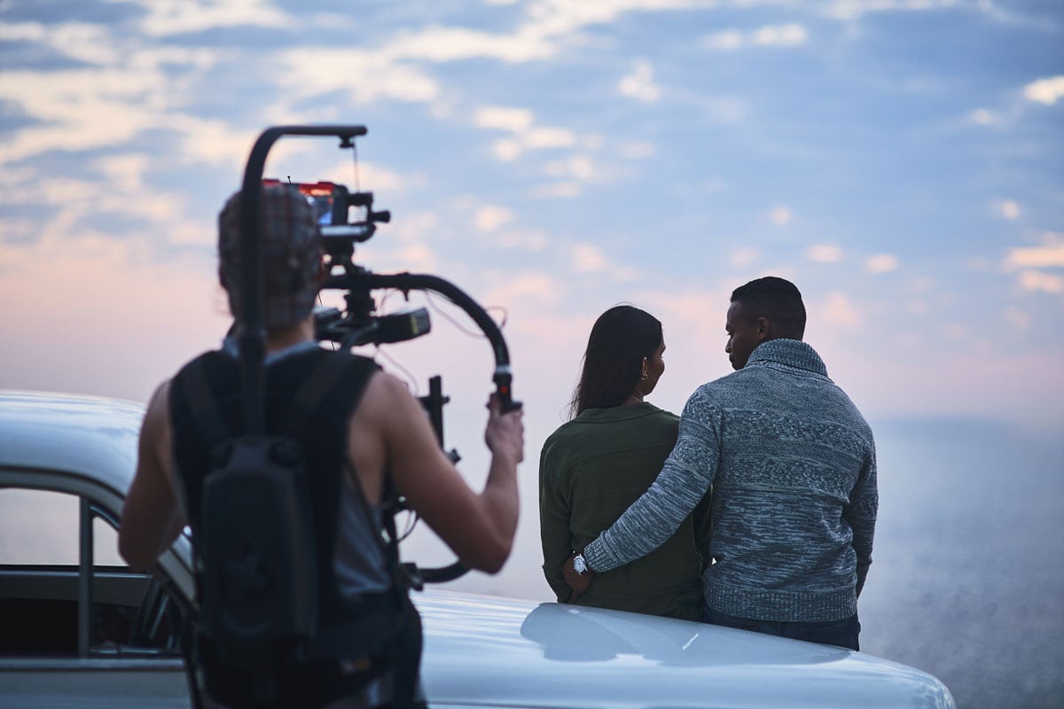 People filming a romantic scene with a camera on a tripod during sunset.