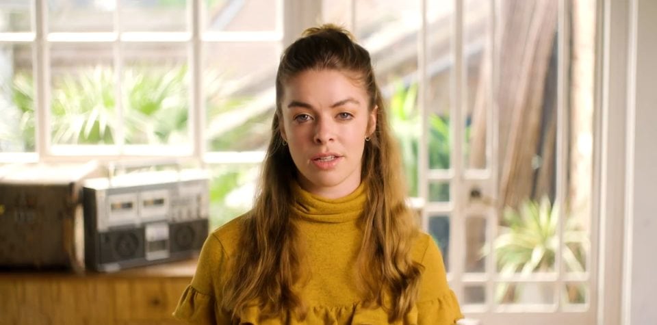 Young woman with long wavy hair in a yellow top speaking in a bright, natural light-filled room.
