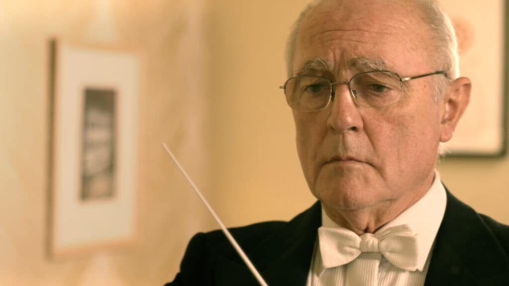 Distinguished senior conductor in tuxedo and glasses, focusing on a musical score with a baton.