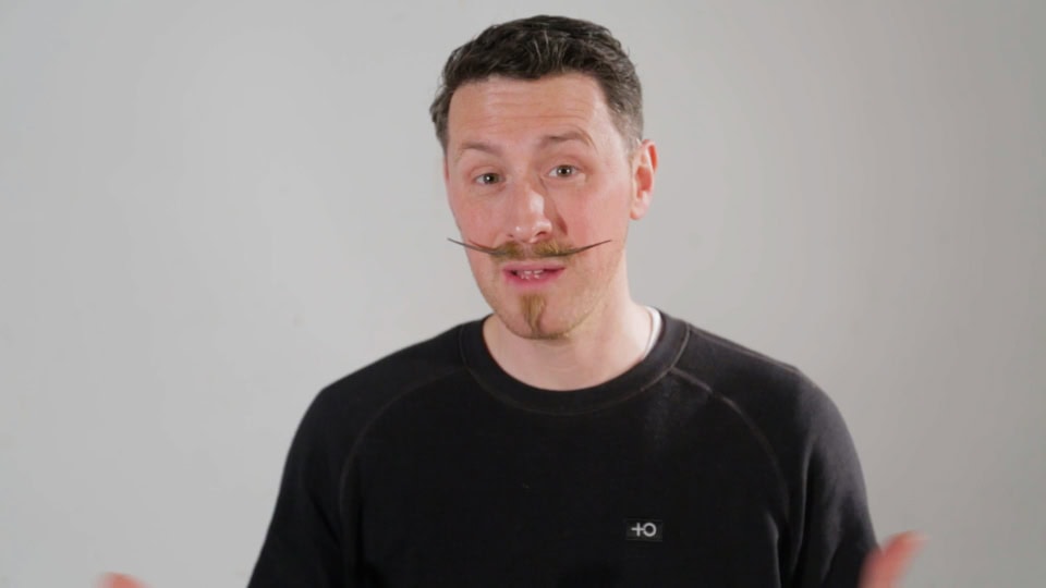 Vibrant man with styled moustache and beard, speaking against a plain grey background.