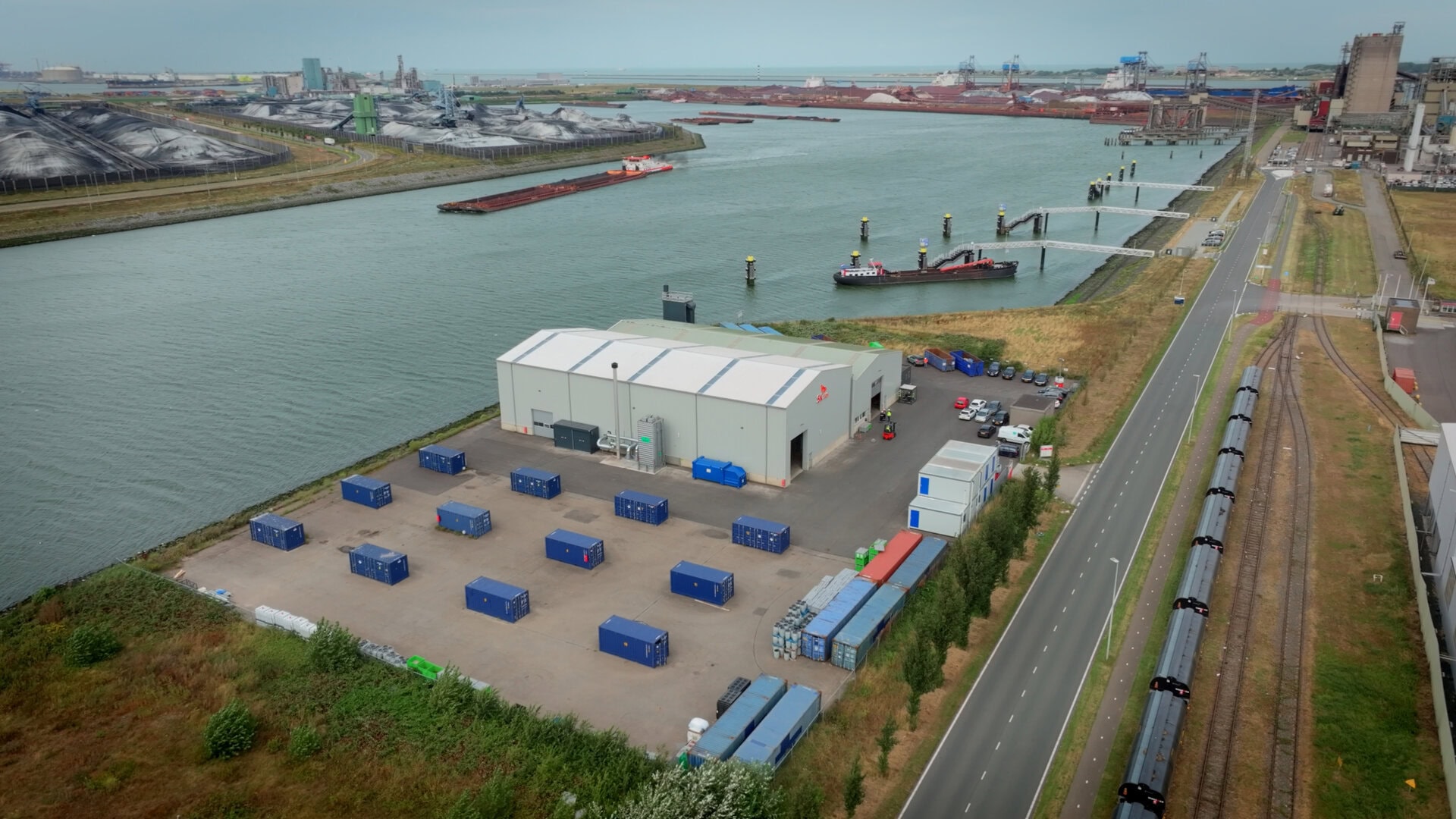 Industrial warehouse and shipping containers along a river with cargo ships and port infrastructure.