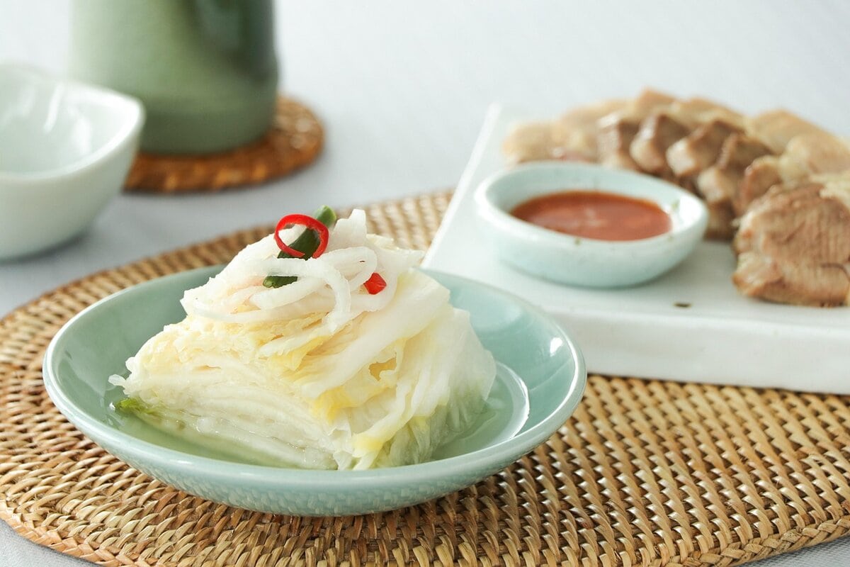 A tempting close-up photograph of a classic Korean meal: Baek-kimchi (white, non-spicy Kimchi) and Suyuk (boiled pork slices). On a rustic wooden table in a cozy Hanok-style restaurant, a clear and refreshing plate of Baek-kimchi is beautifully arranged, featuring crunchy napa cabbage slices with julienned vegetables and red chili threads. Adjacent to it, tender, glistening Suyuk pork belly slices are perfectly arranged, emitting gentle steam. The dish is accompanied by small bowls of Ssamjang (soybean dipping sauce), Saeu-jeot (salted shrimp), fresh garlic slices, green chilies, and sweet-and-spicy dried radish strips (Mumallaengi). A set of chopsticks is captured in motion, about to wrap a slice of Suyuk in a piece of Baek-kimchi. Natural daylight illuminates the textures and colors. This image embodies the authentic, heartwarming taste of Korean cuisine and the perfect pairing of flavors. The camera angle is an eye-level close-up, focusing sharply on the food.