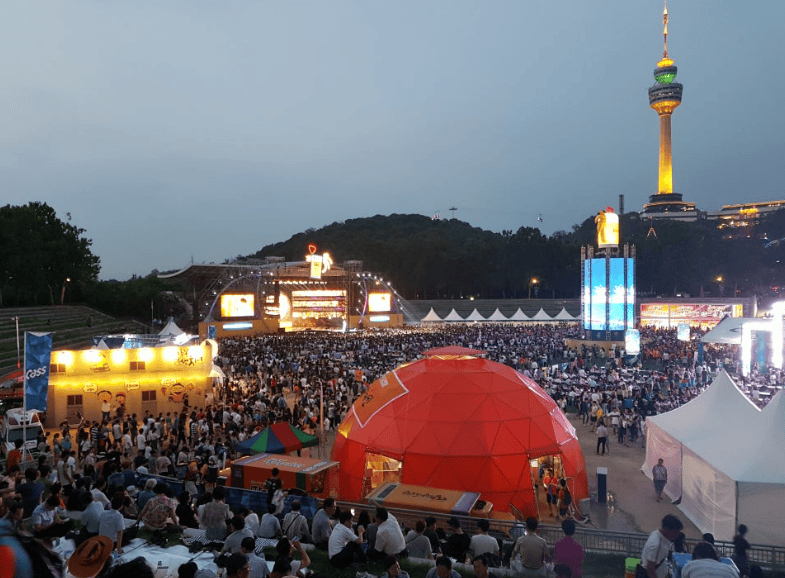 Lively crowd celebrating at the Daegu Chimaek Festival with fried chicken and beer under vibrant stage lights.