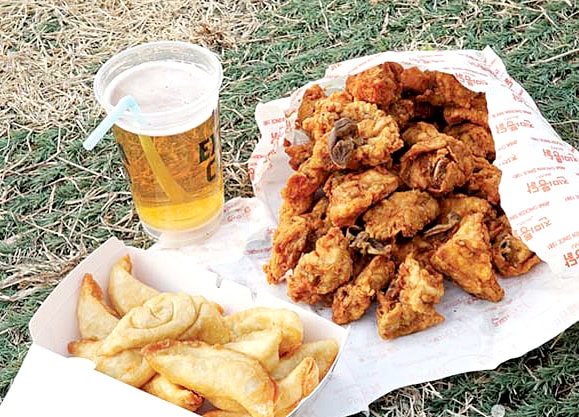 A relaxing Chimaek picnic on a grassy field with crispy fried chicken, a tall glass of draft beer, and crunchy potato chips on a mat.